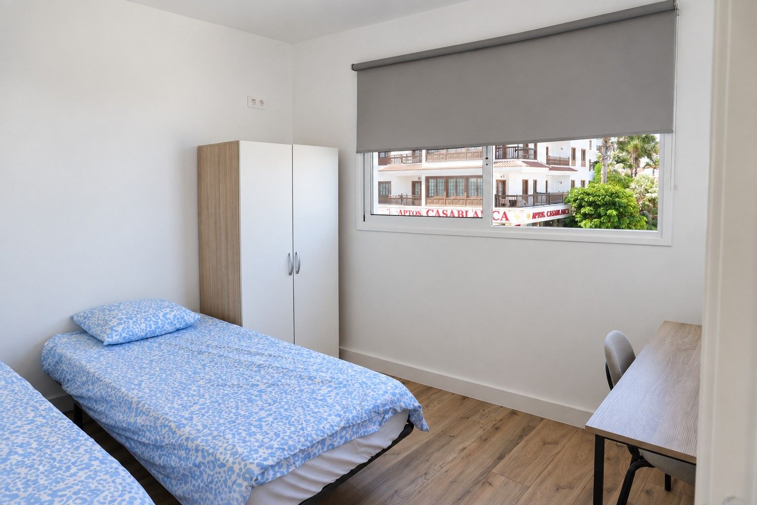 Private room with two single beds in blue bedding, a wardrobe, a desk and a large window onto the street