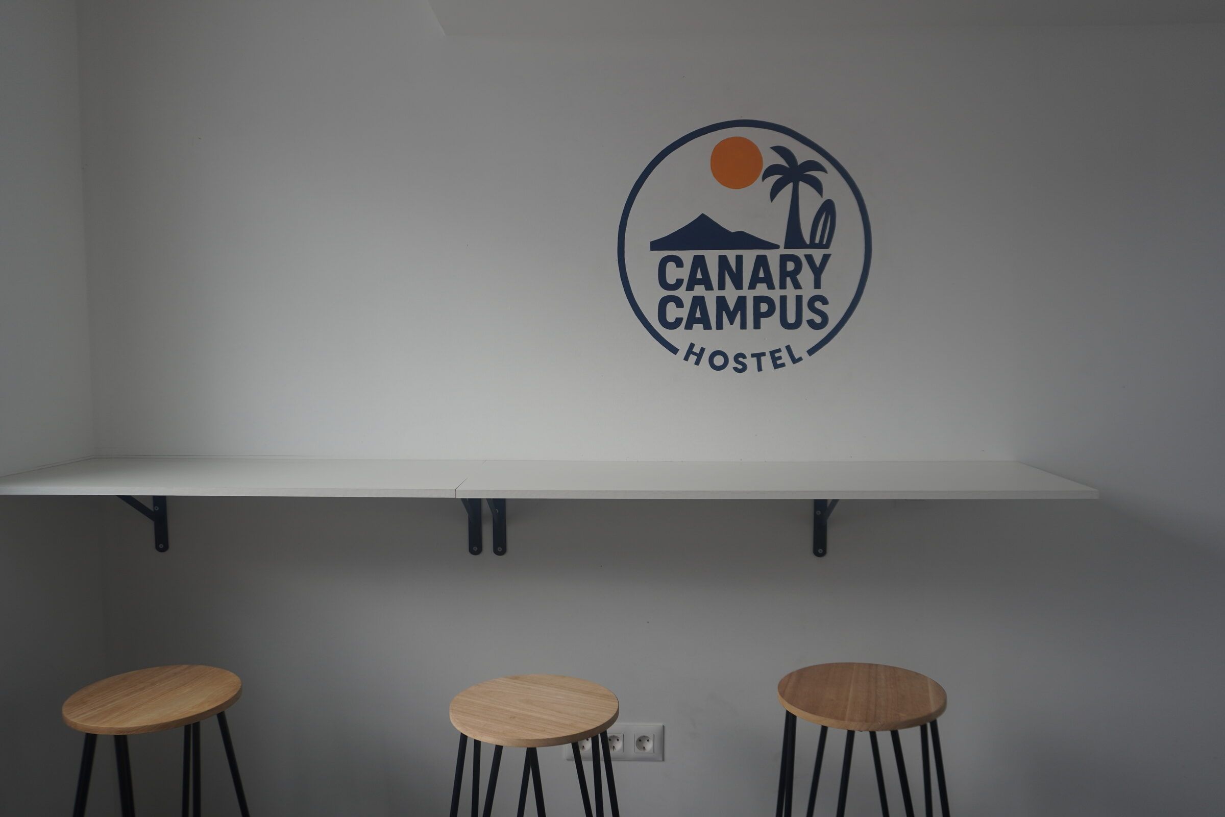 Breakfast bar with three wooden stools under the Canary Campus Hostel logo