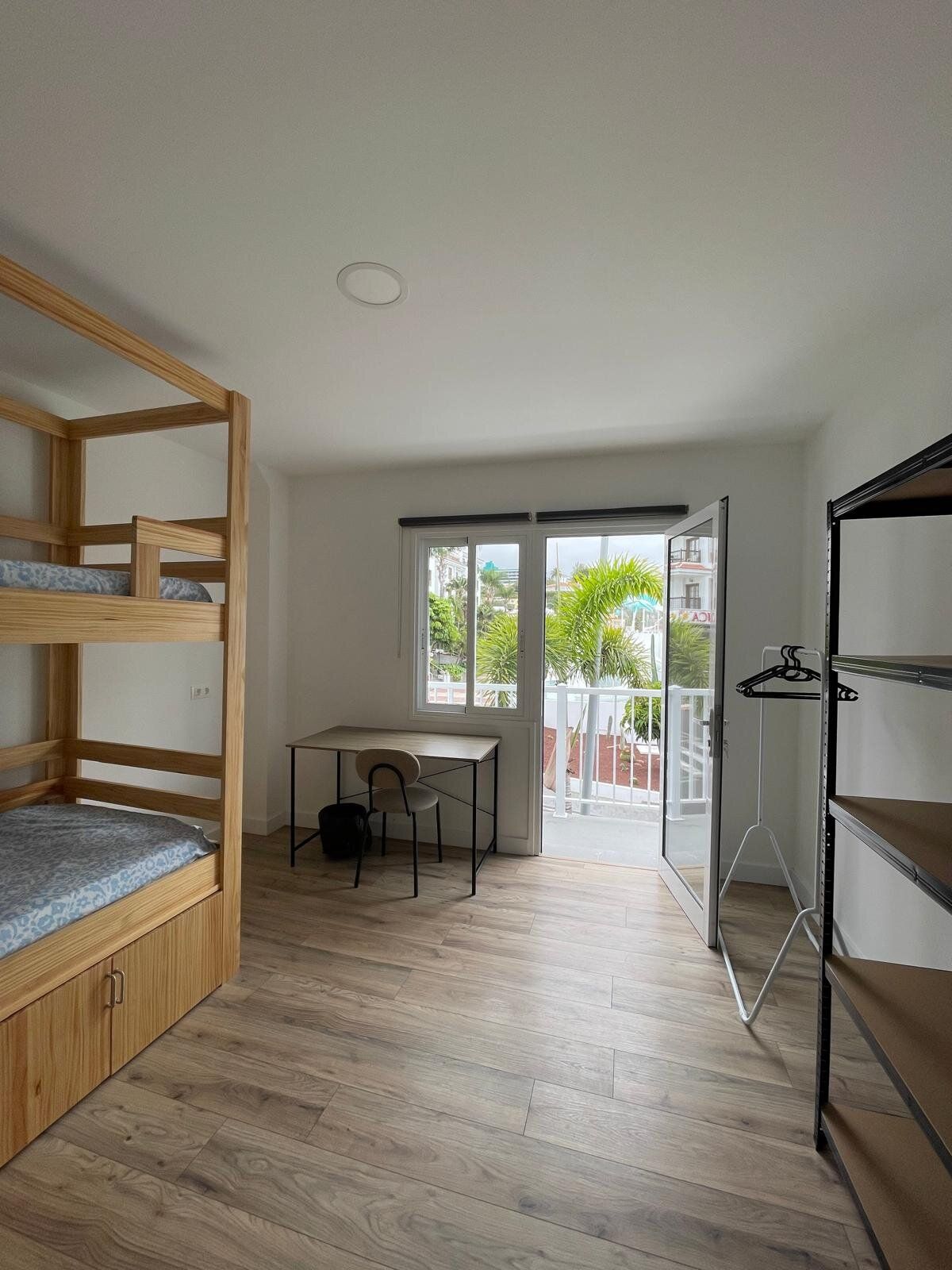 Bunk bed, desk and shelving in a room with an open balcony door and palm trees outside